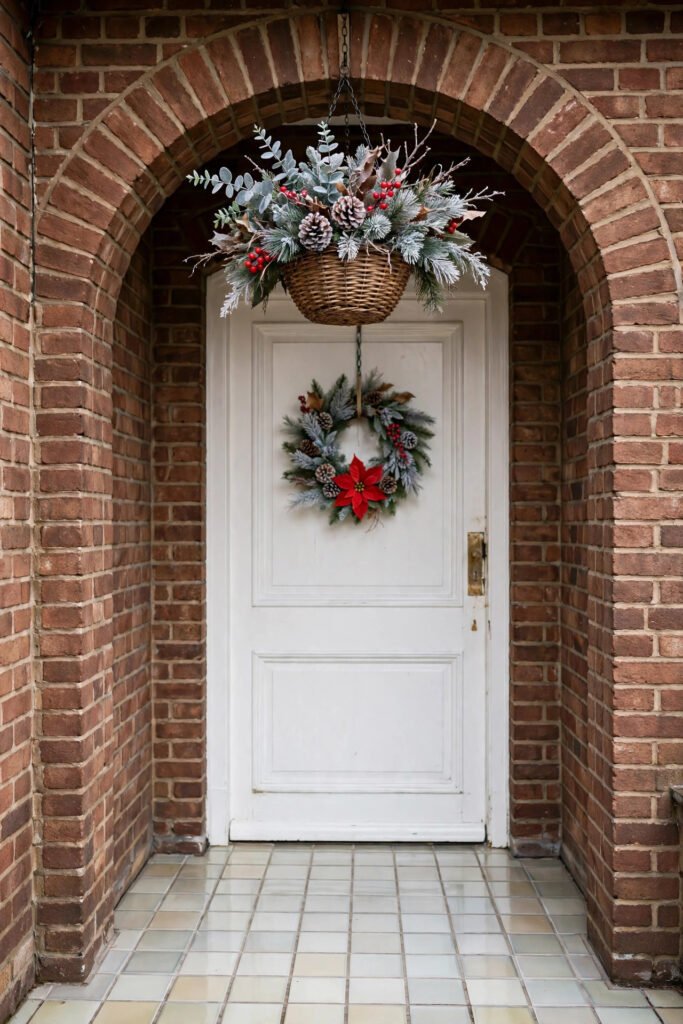 Arched brick entry with wicker hanging basket of frosted greens, pinecones, red berries and twigs, small wreath with red poinsettia on white door, clear tiled entry